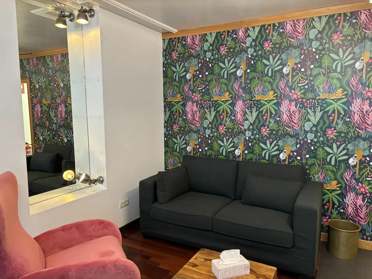 Zona de conversación de la consulta: sofá gris oscuro ante una pared con papel pintado botánico, butaca de terciopelo rosa y espejo con focos de luz cálida